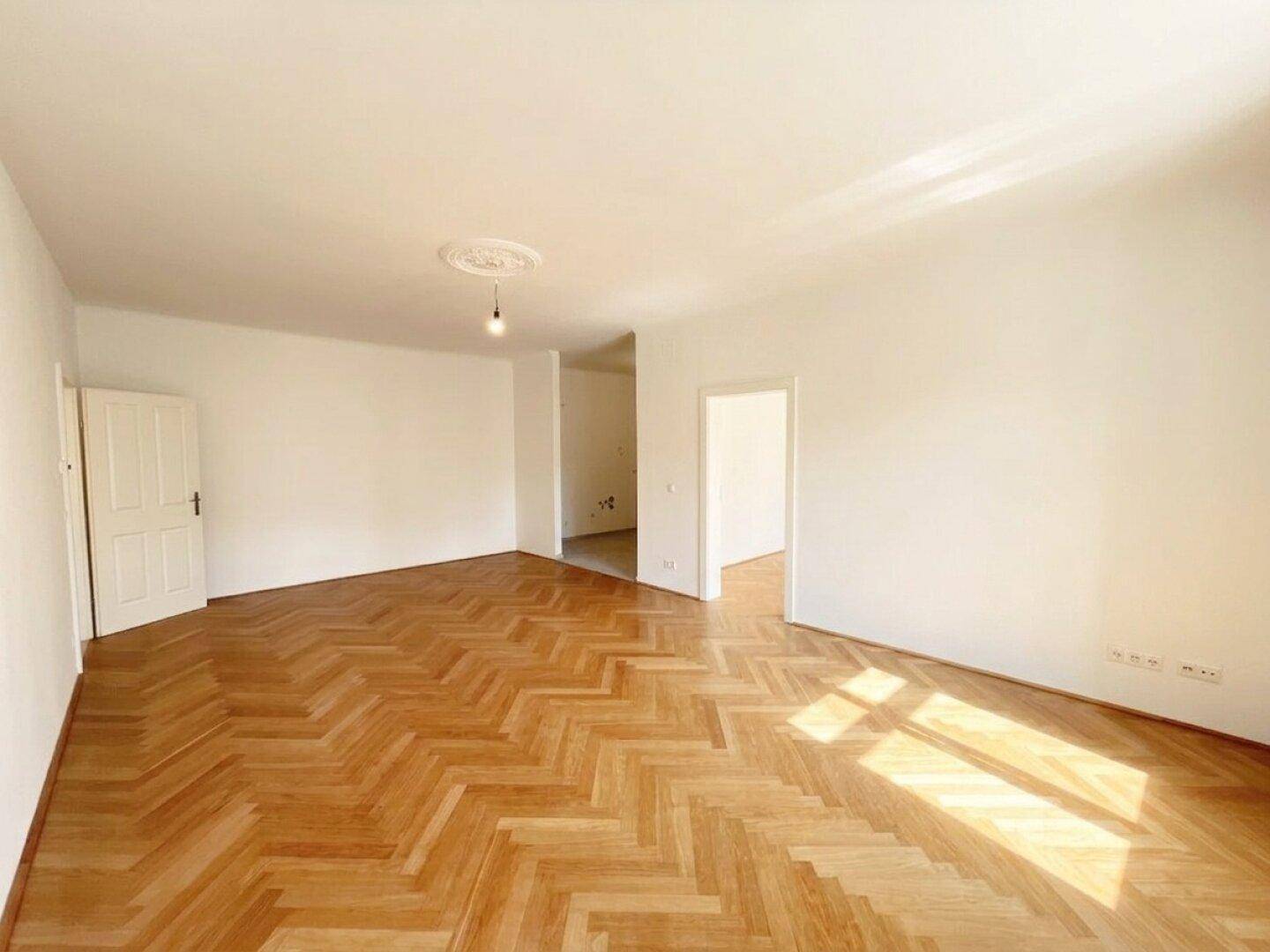Großzügiger Wohnbereich mit Fischgrätparkett und weiß gestrichenen Wänden, bereit zur individuellen Möblierung.