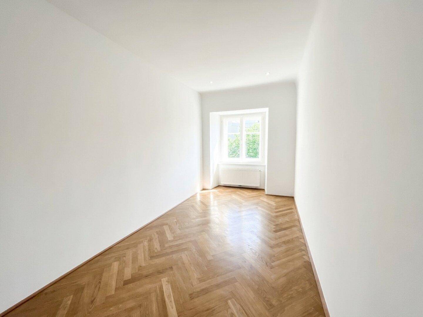 Lichtdurchflutetes Schlafzimmer mit Fischgrätparkett und großem Fenster, das viel Helligkeit in den Raum bringt.
