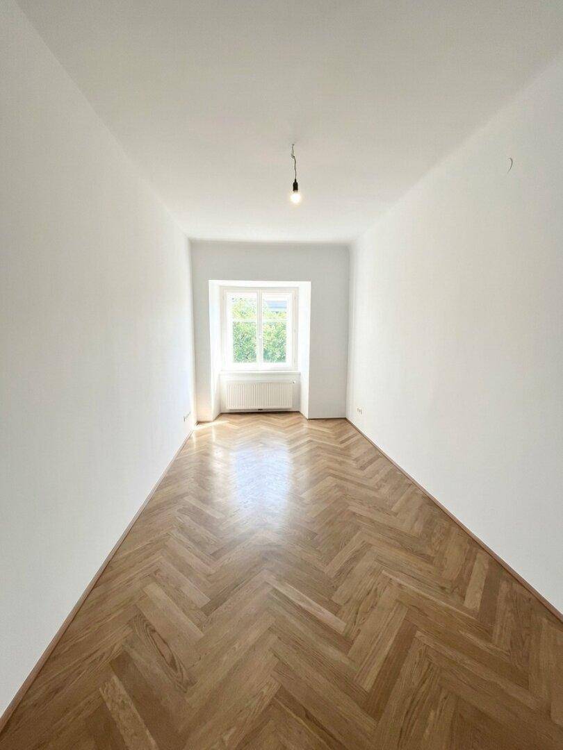 Leeres Schlafzimmer mit Fischgrätparkett und einem Fenster, das den Raum mit angenehmem Tageslicht füllt.