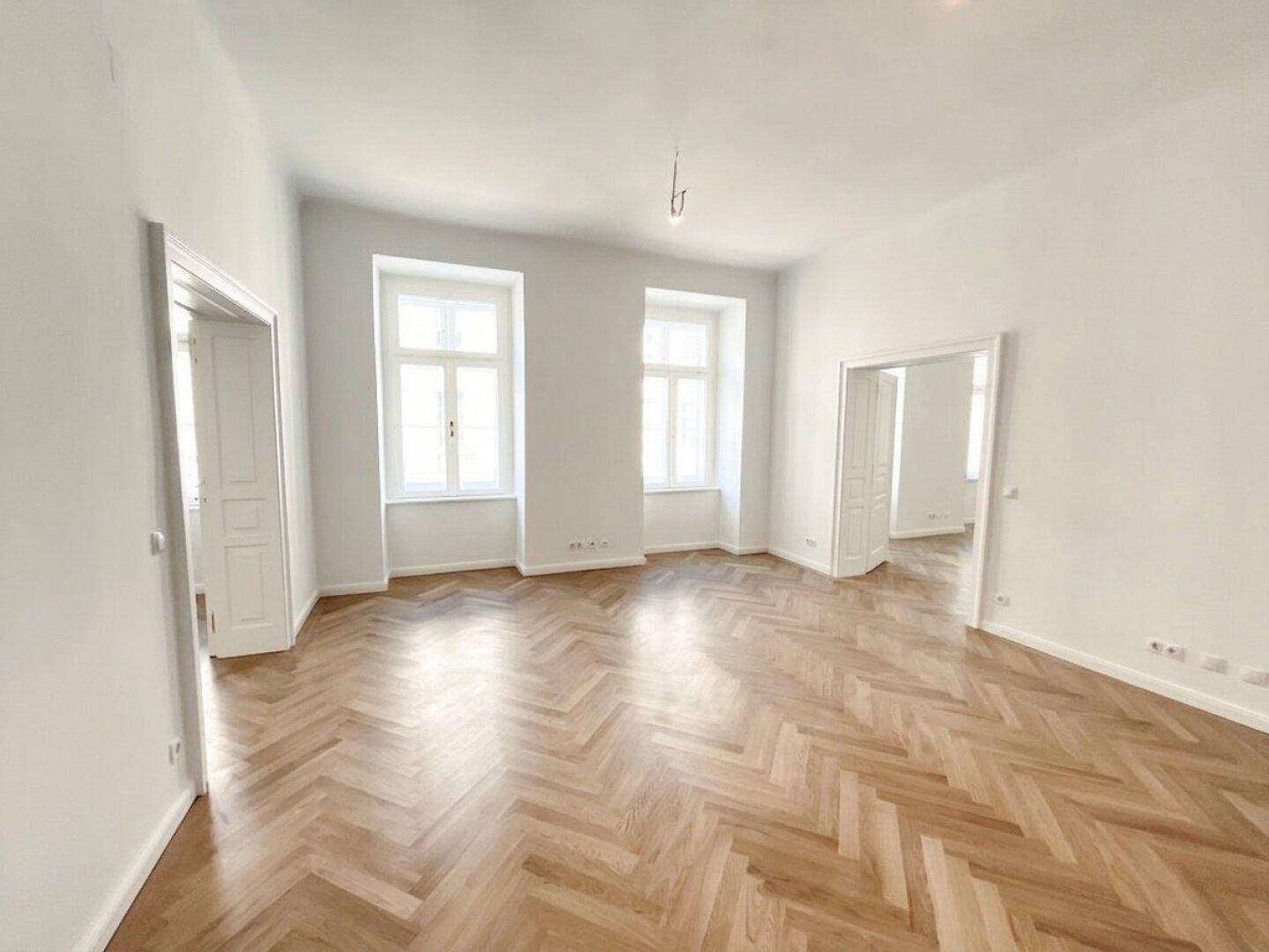 Großes, helles Zimmer mit Fischgrätparkett und zwei hohen Fenstern, ideal für Wohn- oder Schlafbereich.