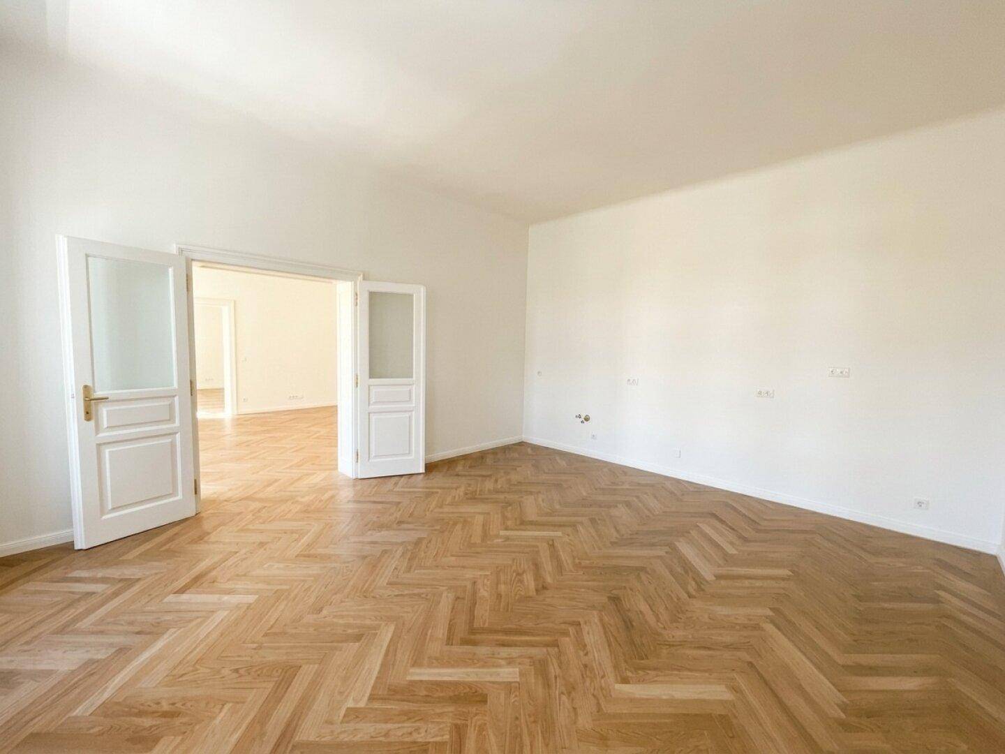 Geräumiges Zimmer mit Fischgrätparkett und Doppeltüren, die zu einem weiteren Raum führen.