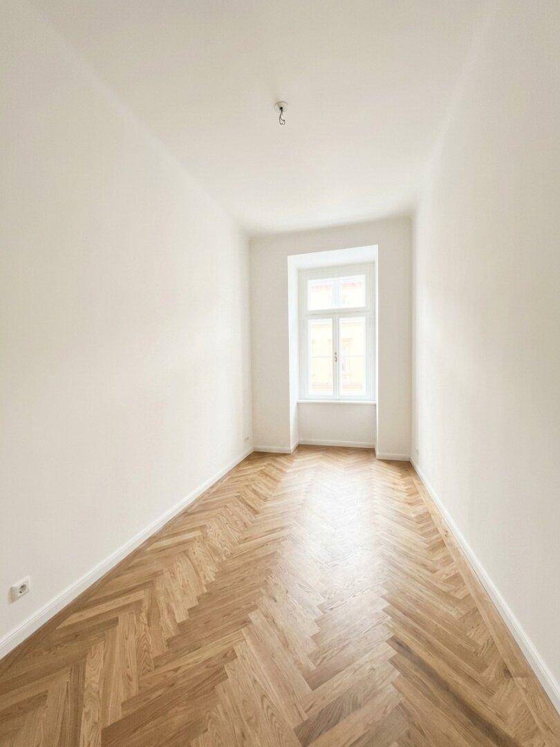 Gemütliches Zimmer mit Fischgrätparkett und einem hohen Fenster, ideal als Schlaf- oder Arbeitszimmer.