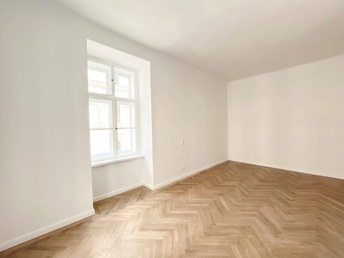 Lichtdurchflutetes Zimmer mit Fischgrätparkett und einem großen Fenster, das viel Tageslicht hereinlässt.
