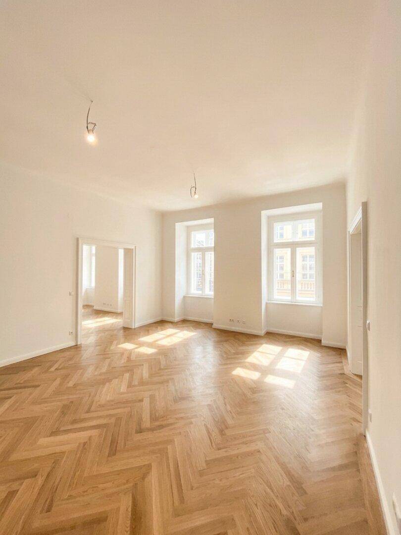 Lichtdurchflutetes Zimmer mit Fischgrätparkett und hohen Fenstern, das eine einladende Atmosphäre bietet.