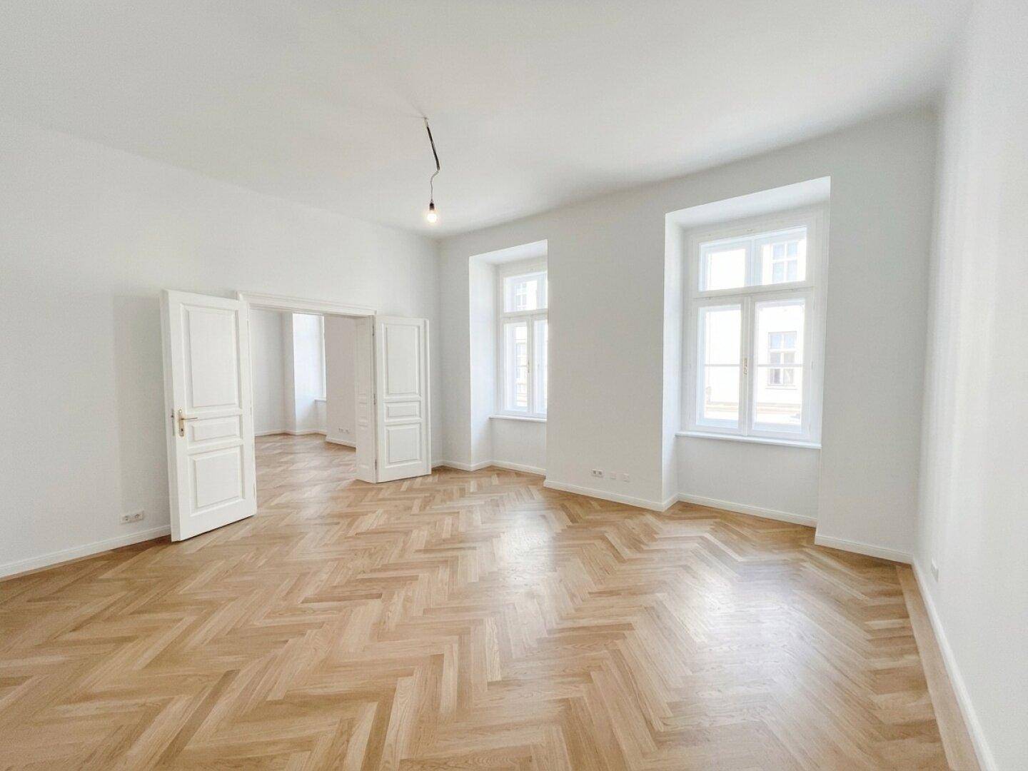Geräumiger, heller Raum mit Fischgrätparkett und hohen Fenstern, ideal für Wohn- oder Essbereich.
