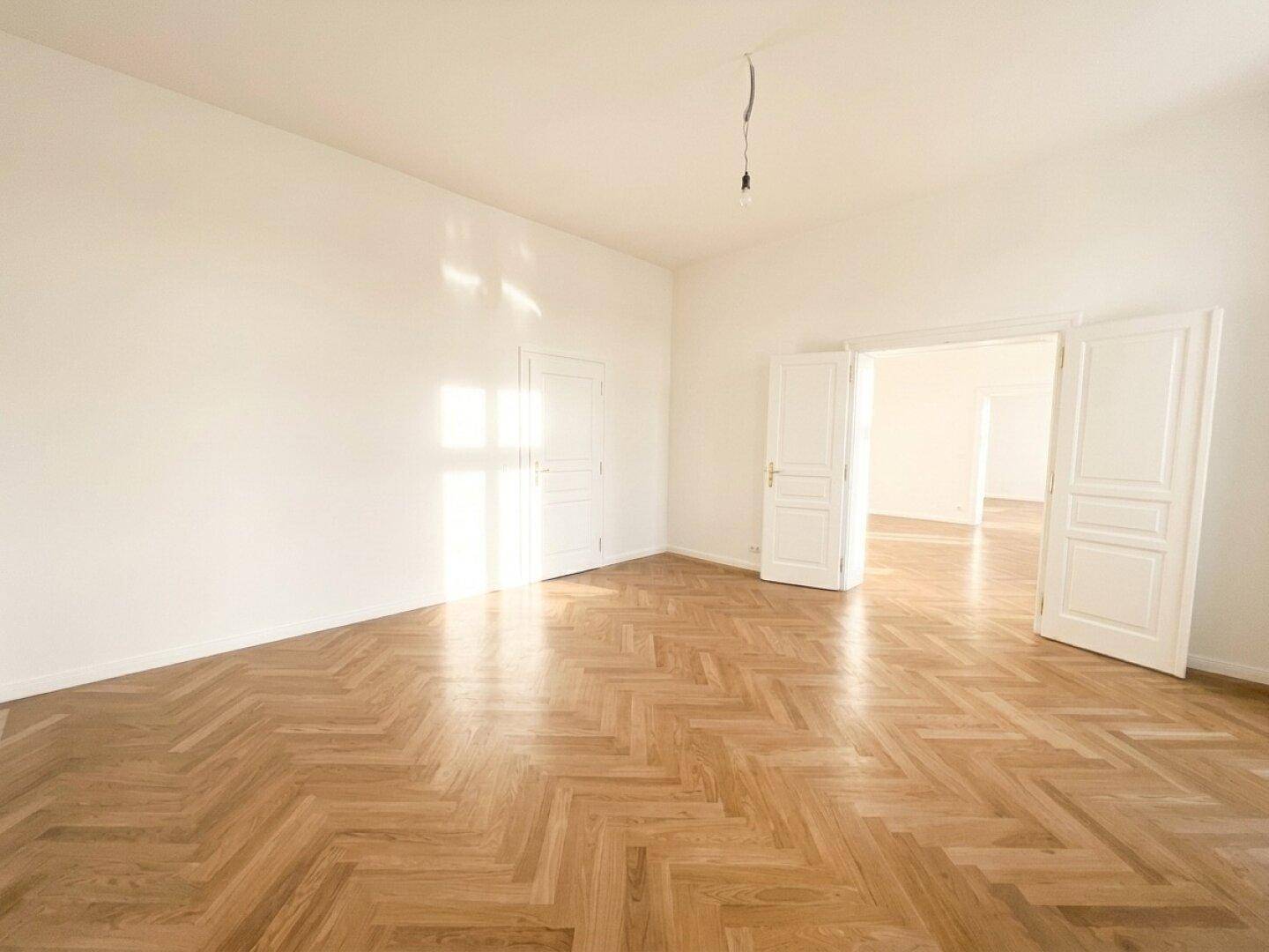 Geräumiger, heller Raum mit Fischgrätparkett und hohen Fenstern, ideal für Wohn- oder Essbereich.