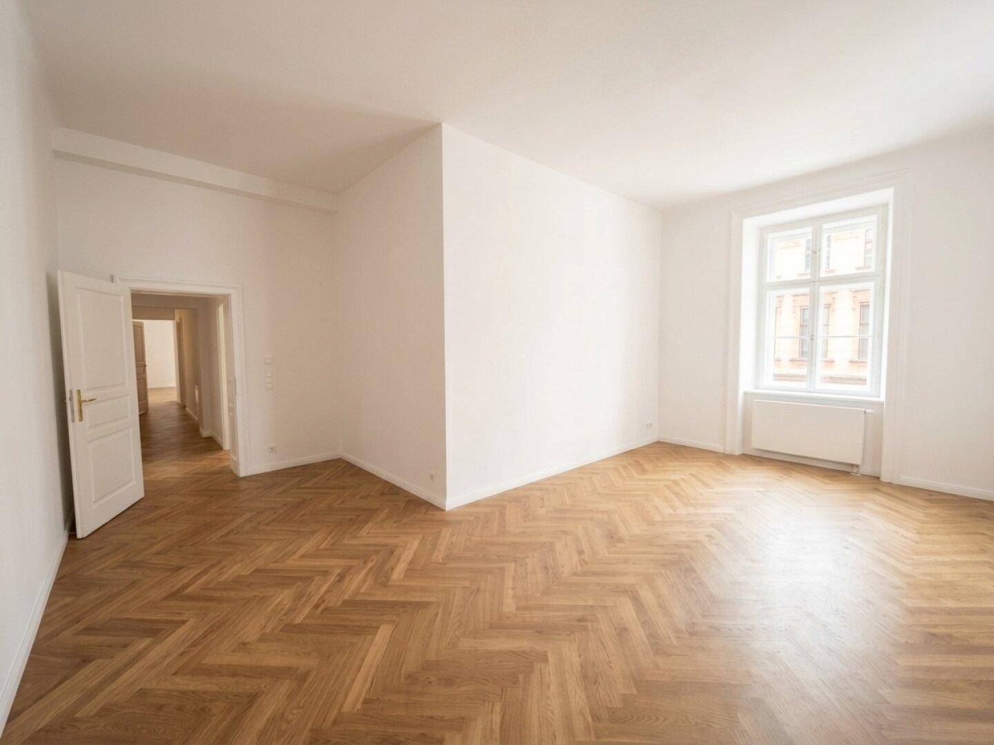 Heller Raum mit Fischgrätparkett und einem großen Fenster, das viel Tageslicht hereinlässt.