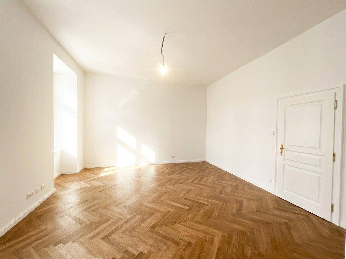 Helles Wohnzimmer mit Fischgrätparkett und einem großen Fenster, das viel Licht hereinlässt und den Raum erhellt.
