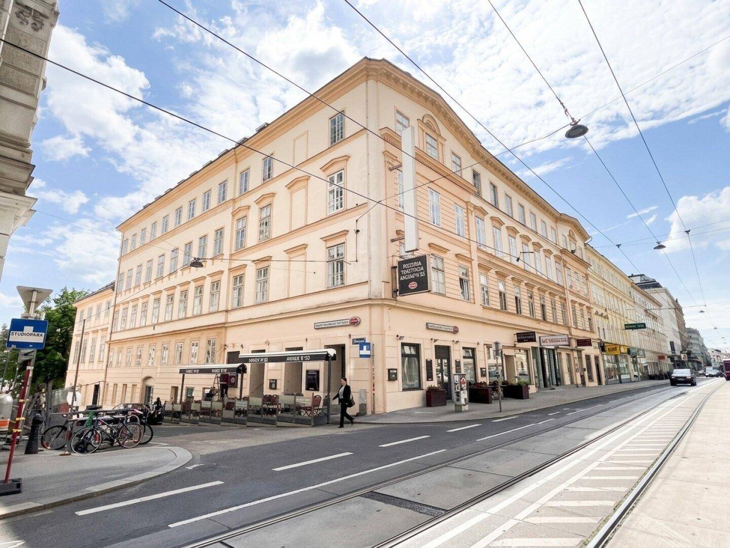 Großes Eckgebäude an einer belebten Straße mit Geschäften und Außengastronomie unter blauem Himmel.