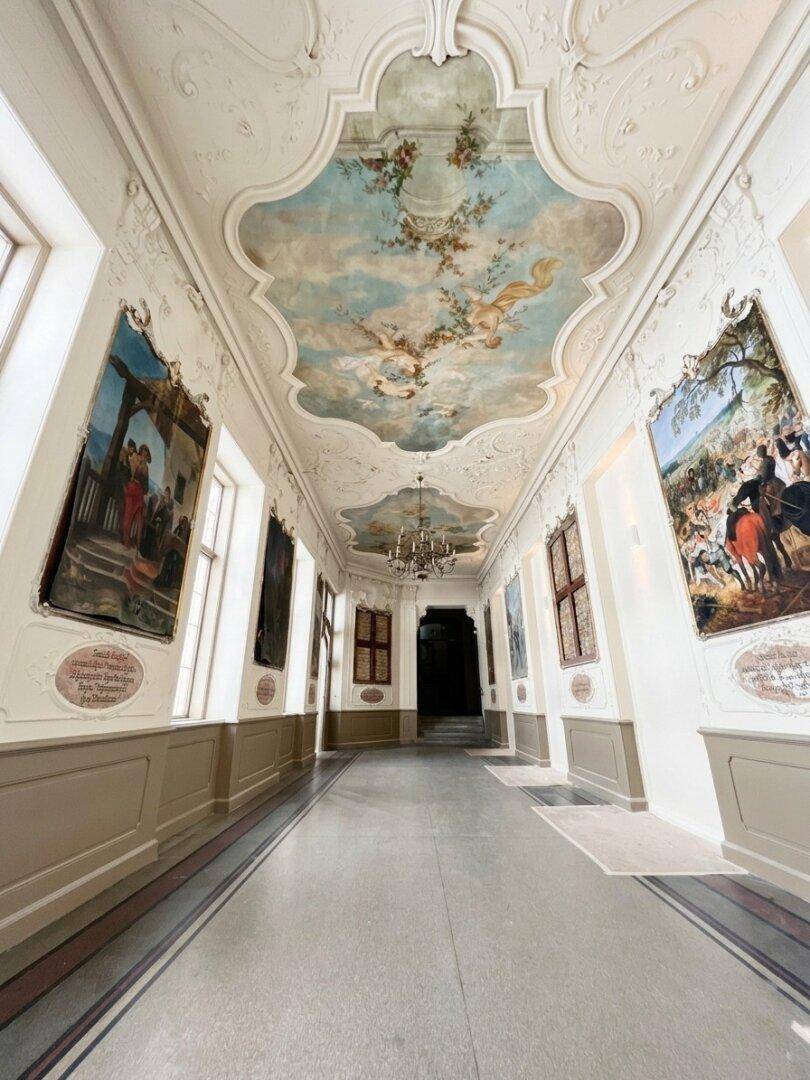Prächtiger Flur mit kunstvoll bemalter Decke, großen Gemälden und hohen Fenstern in einem historischen Gebäude.