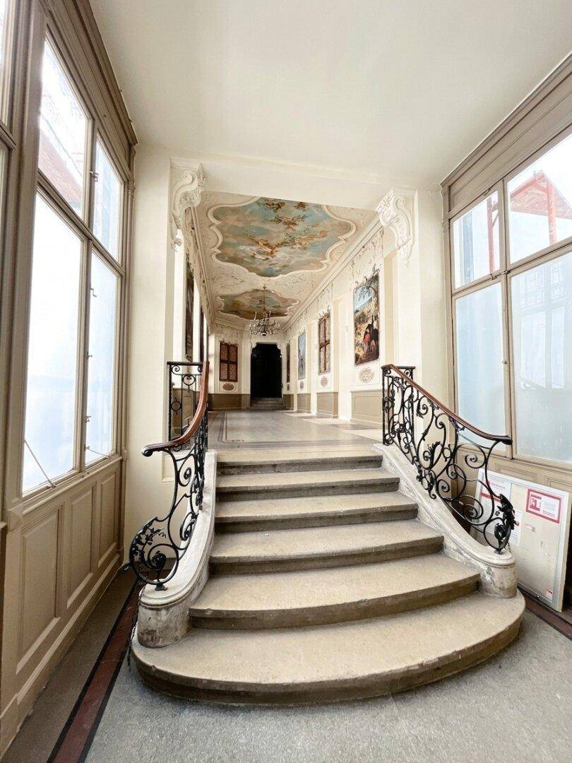 Elegantes Treppenhaus mit kunstvollen Geländern, bemalter Decke und großen Fenstern, die viel Licht hereinlassen.
