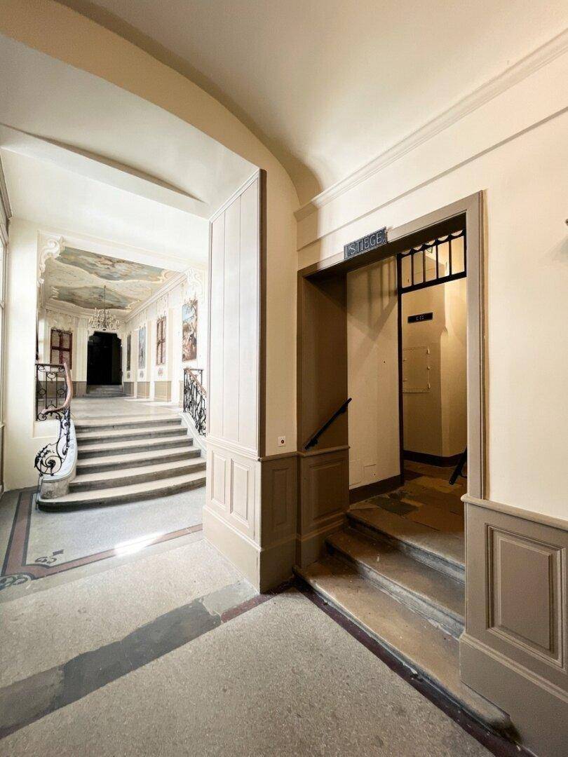 Historischer Flur mit Zugang zum Aufzug und einer eleganten Treppe im Hintergrund, verziert mit Stuckdecken.