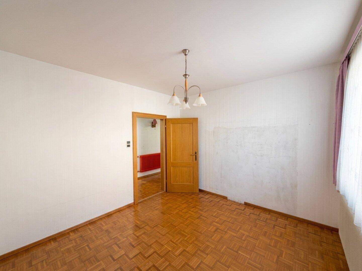 Helles Zimmer mit Parkettboden und einem Fenster mit lila Vorhängen.