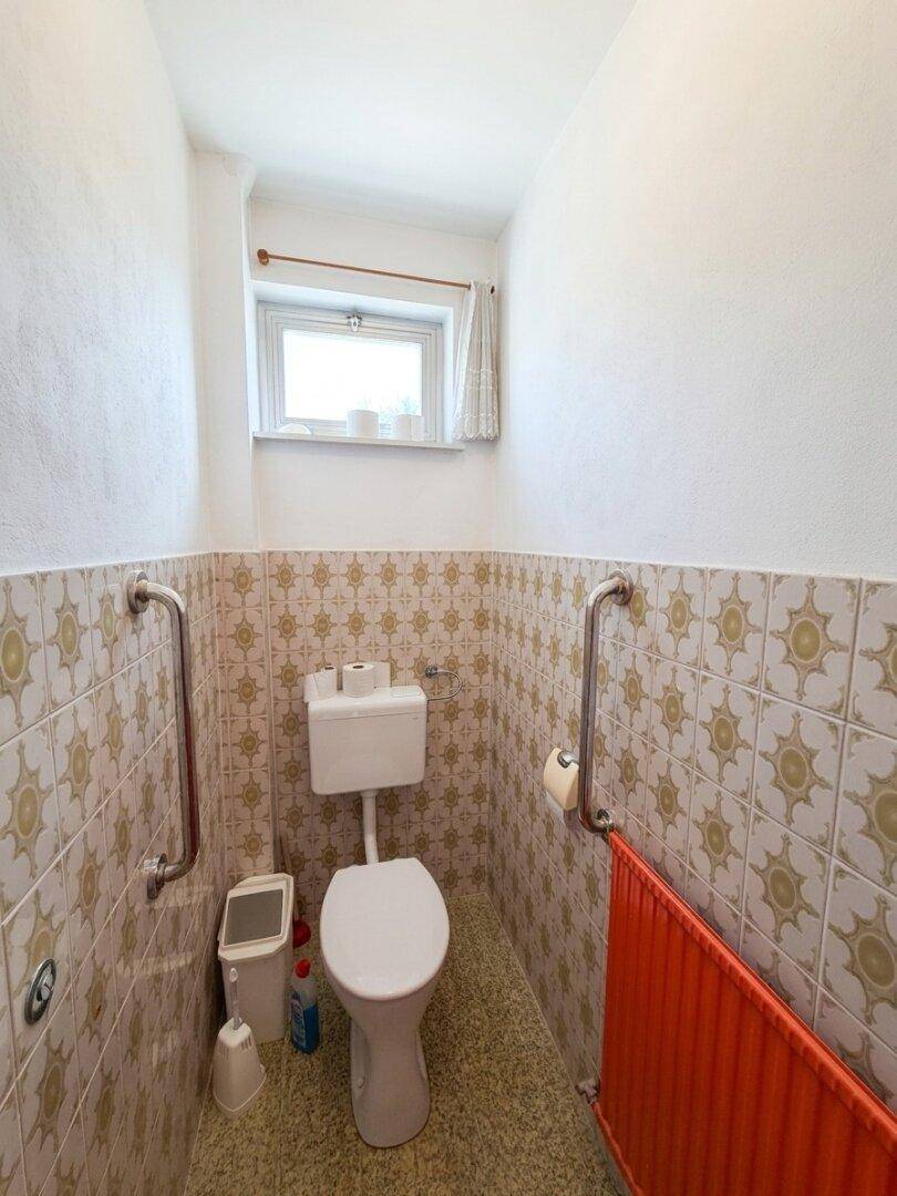 Gäste-WC mit gemusterten Fliesen, Toilette, Fenster und Haltegriffen für Barrierefreiheit.