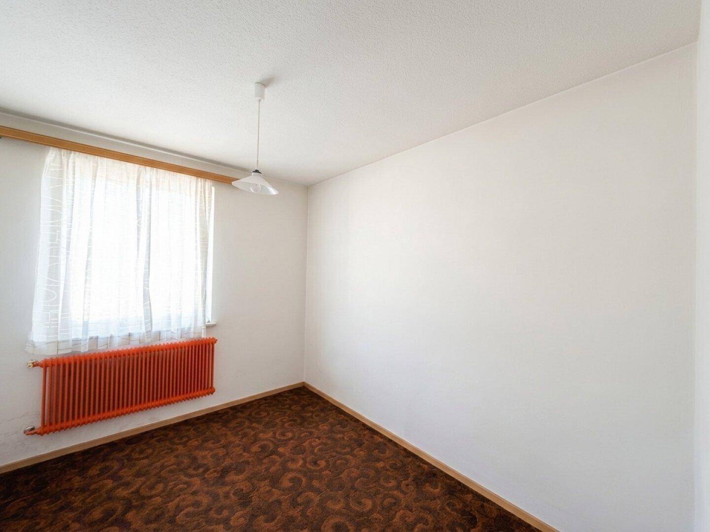 Schlafzimmer mit Teppichboden, weiß gestrichenen Wänden und einem roten Heizkörper.