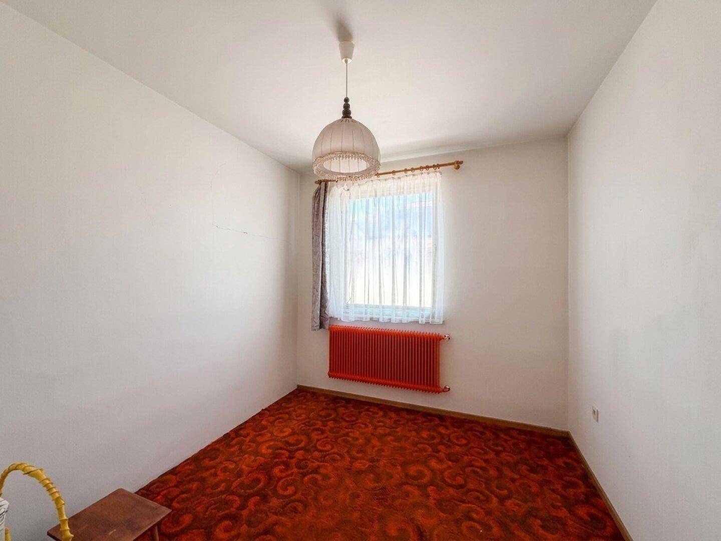 Kleines Schlafzimmer mit gemustertem Teppichboden, rotem Heizkörper und Fenster.