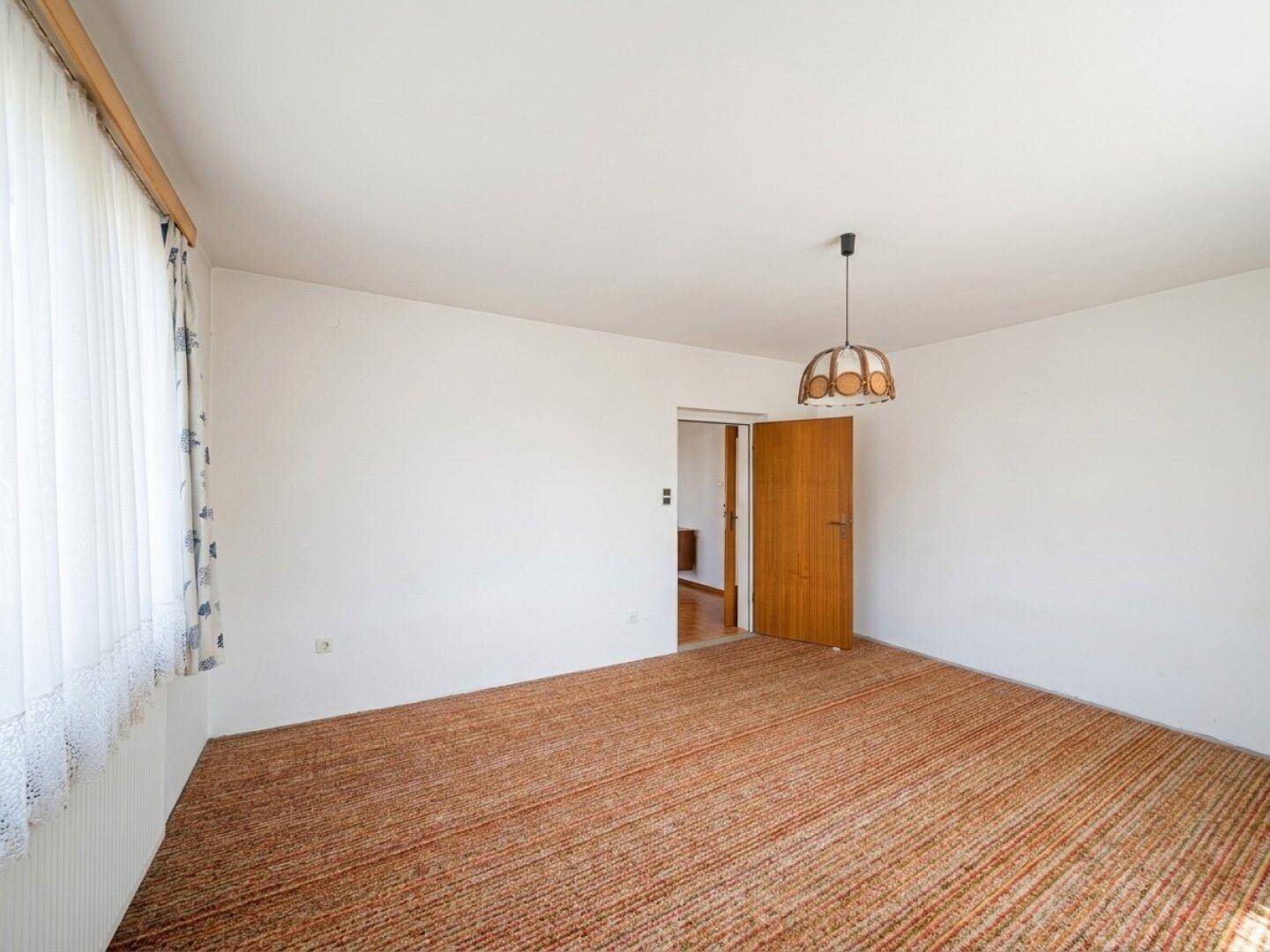 Wohnzimmer mit Teppichboden, Fenster und Zugang zu einem weiteren Raum.