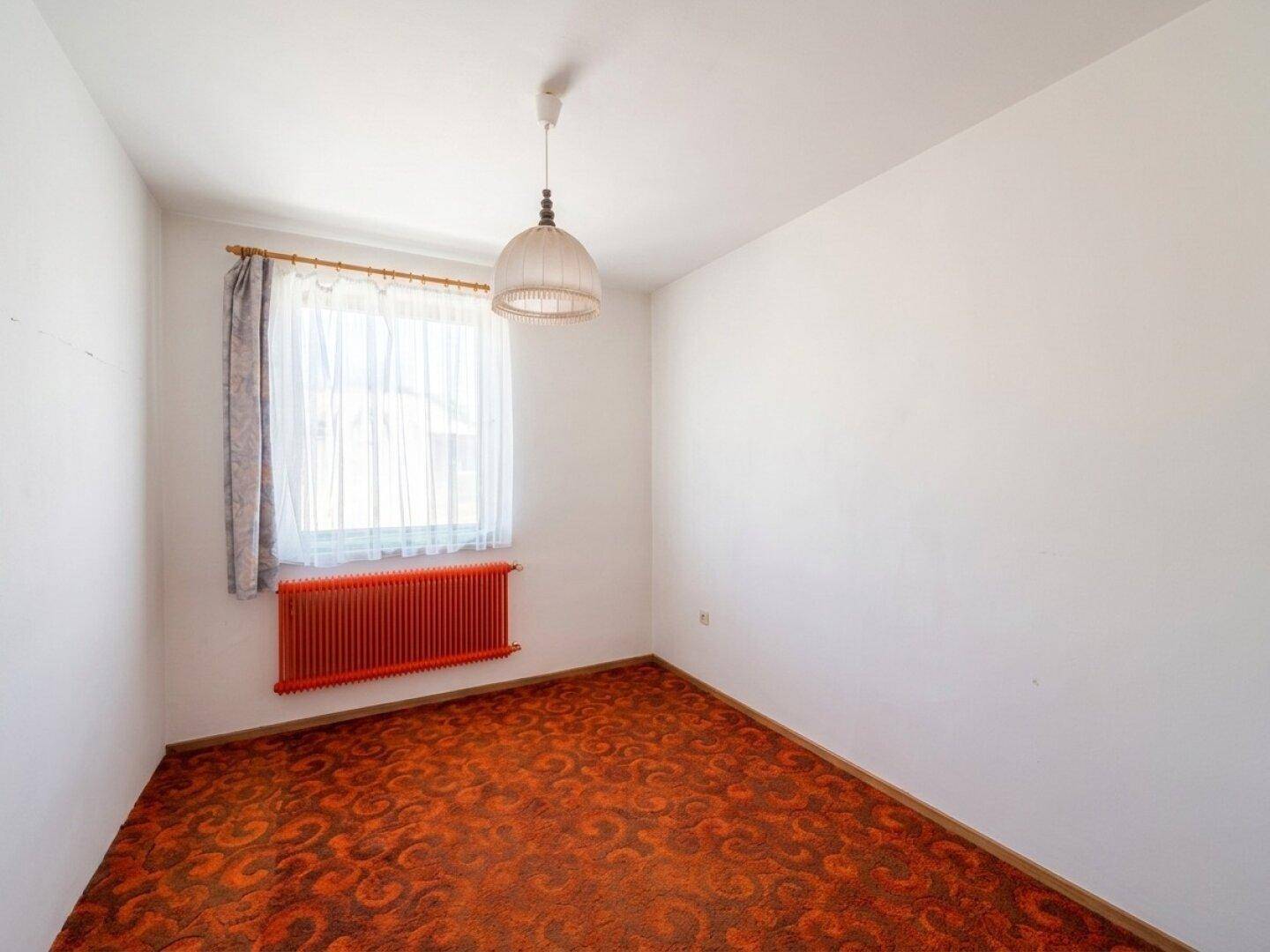 Schlafzimmer mit gemustertem Teppichboden, rotem Heizkörper und Fenster.