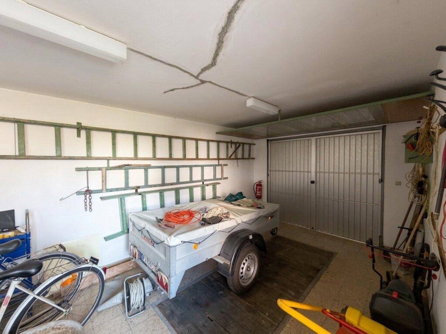 Geräumige Garage mit einem Anhänger, Fahrrädern und Leitern an den Wänden zur Aufbewahrung.