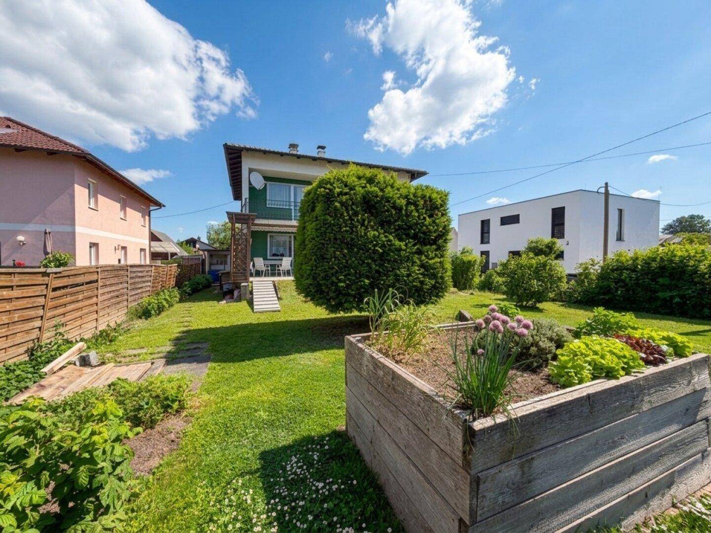 Großer grüner Garten hinter dem Haus mit Hochbeet und Terrasse unter blauem Himmel.