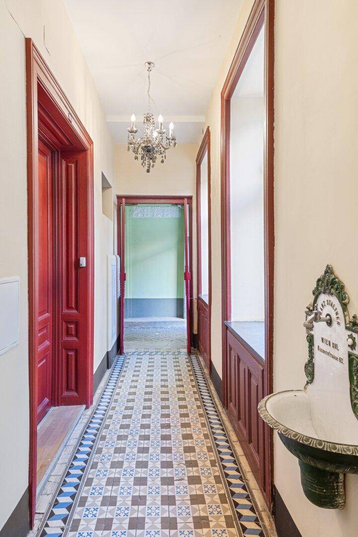 Langer Flur im Altbau mit gemustertem Fliesenboden, roten Türen und einem historischen Waschbecken.