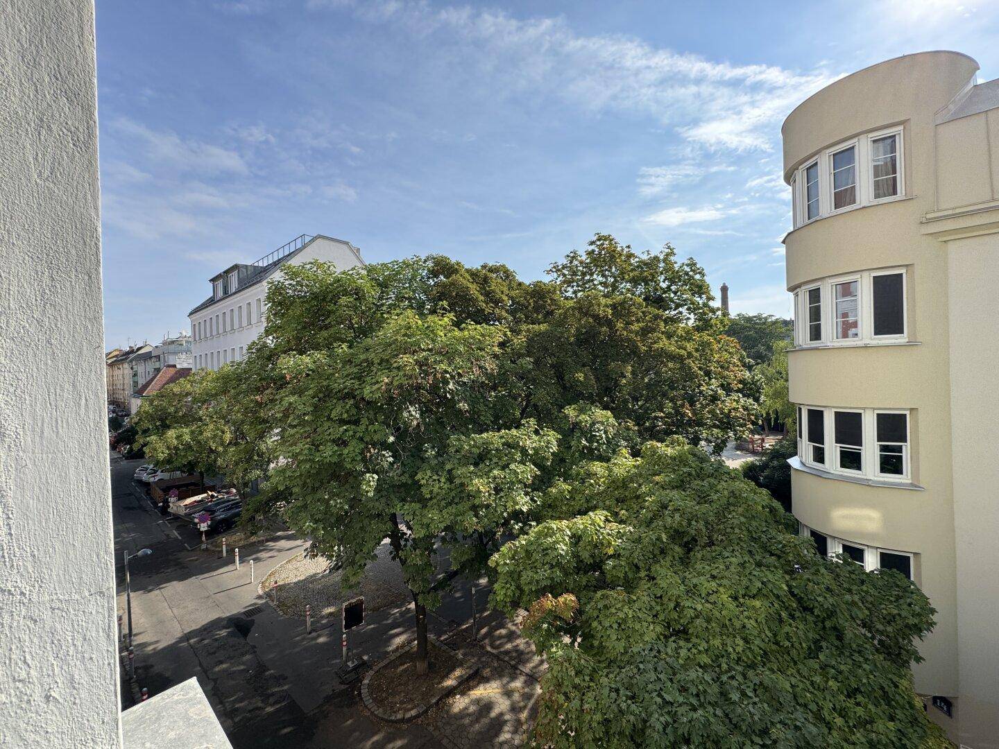 Blick von einem Balkon auf eine grüne Straße mit Bäumen und umliegenden Gebäuden.