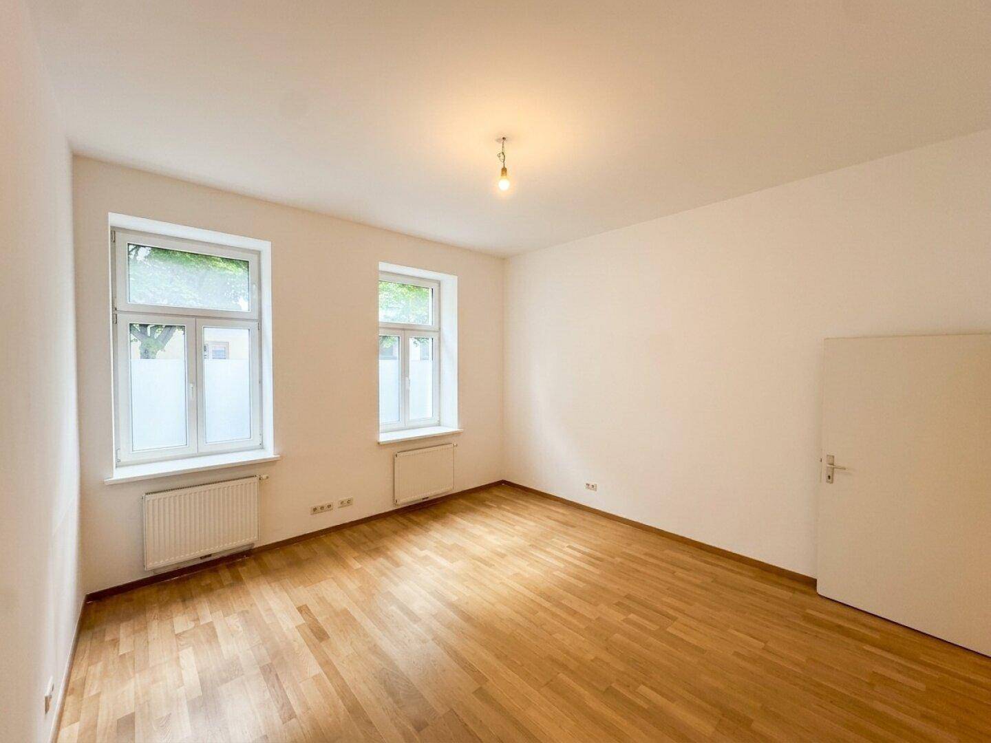 Heller, leerer Raum mit Holzboden und zwei Fenstern, ideal für verschiedene Nutzungen.