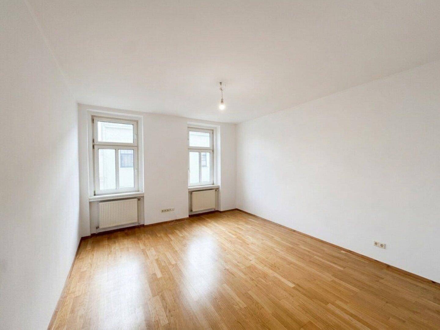 Leeres Zimmer mit Parkettboden und zwei großen Fenstern, bereit zur individuellen Gestaltung.