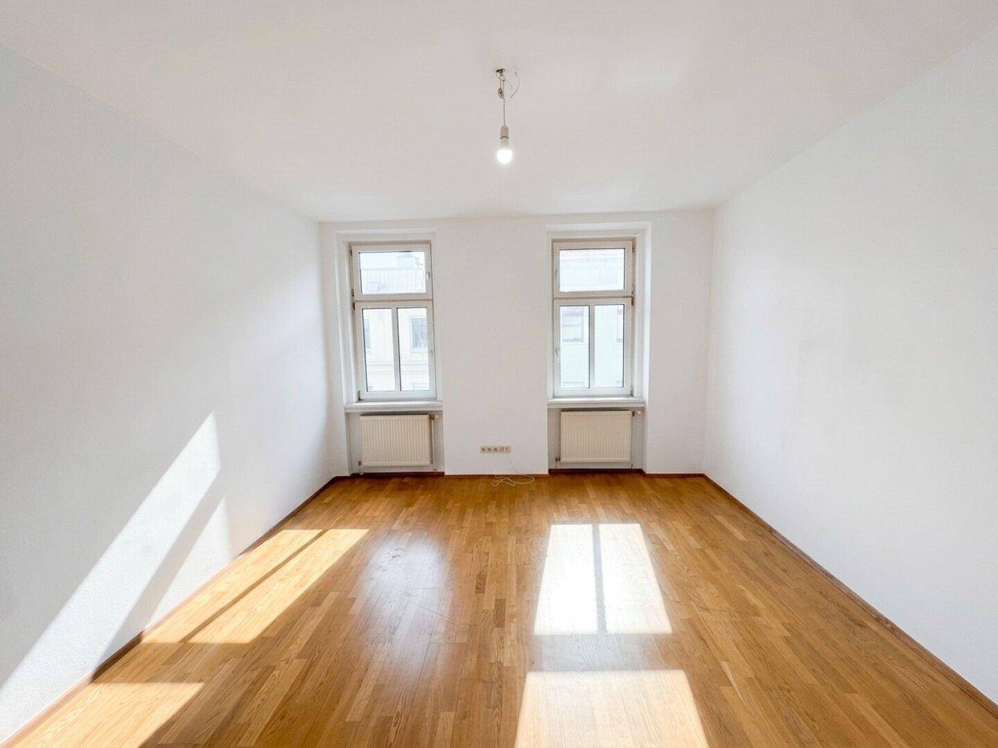 Helles, leeres Zimmer mit Parkettboden und zwei Fenstern, ideal als Schlaf- oder Arbeitszimmer.