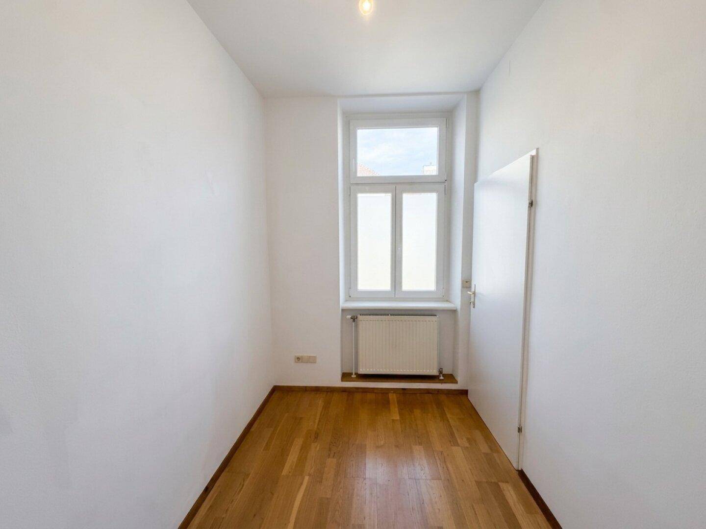 Schmaler Raum mit Parkettboden und hohem Fenster, geeignet als Gäste- oder Ankleidezimmer.