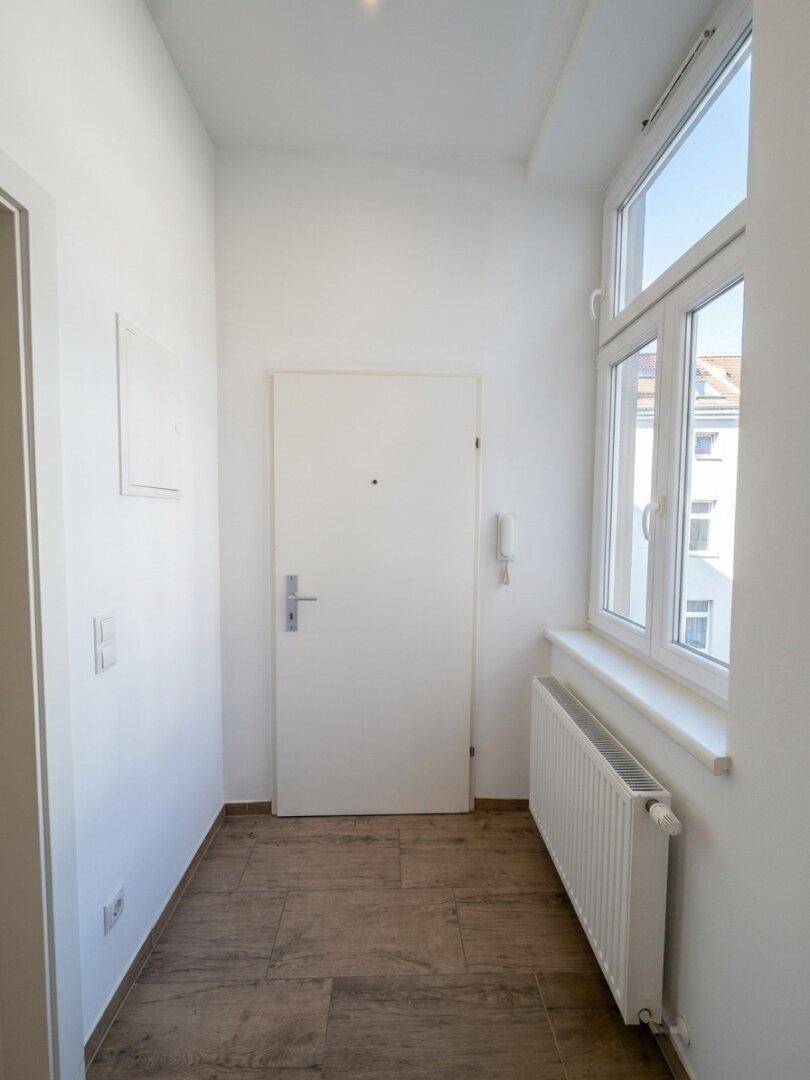 Eingangsbereich mit Fenster, Holzoptik-Fliesenboden und Heizkörper, der viel Tageslicht hereinlässt.