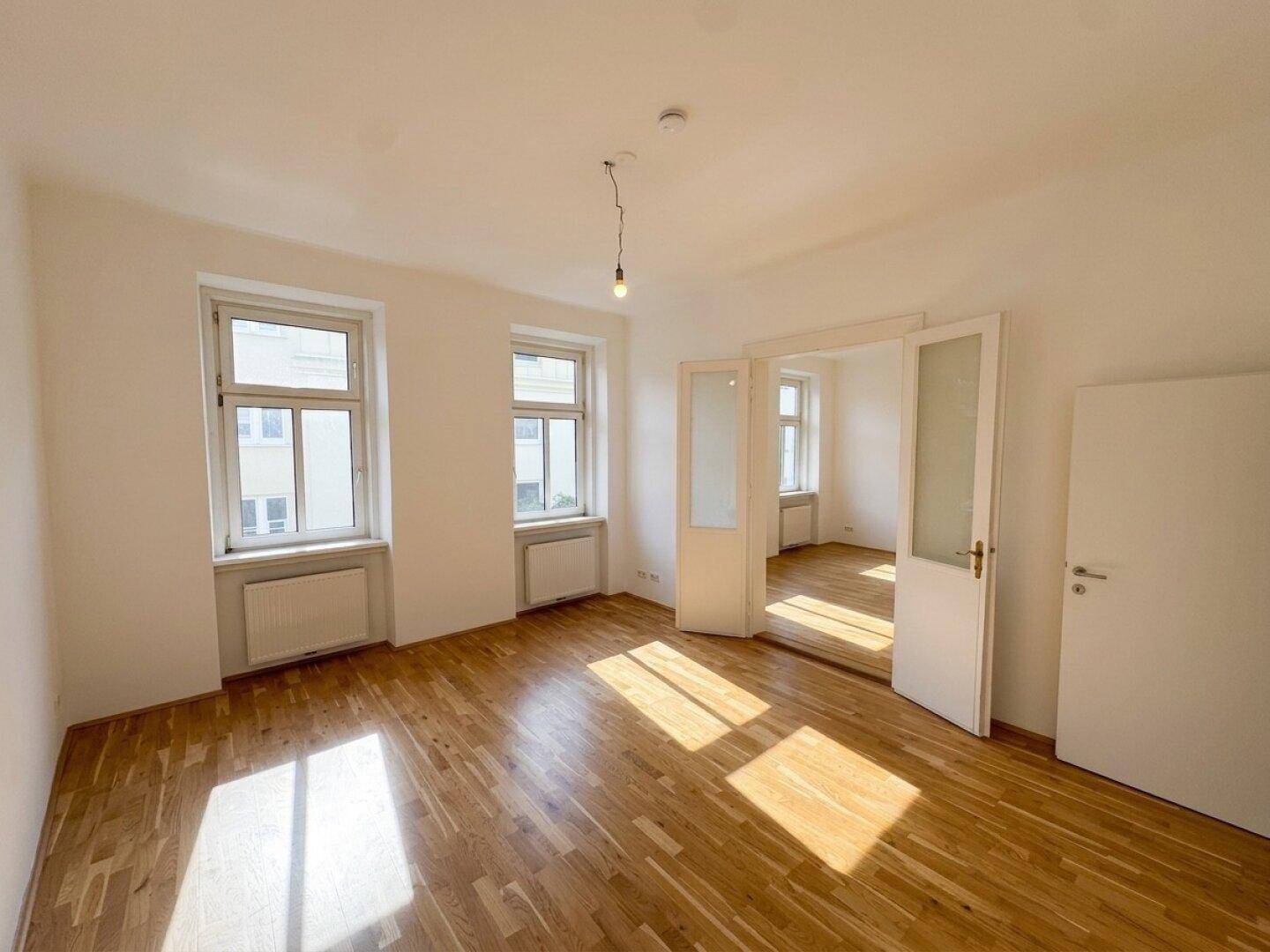 Geräumiges, unmöbliertes Wohnzimmer mit hellem Parkettboden und viel Tageslicht durch große Fenster.