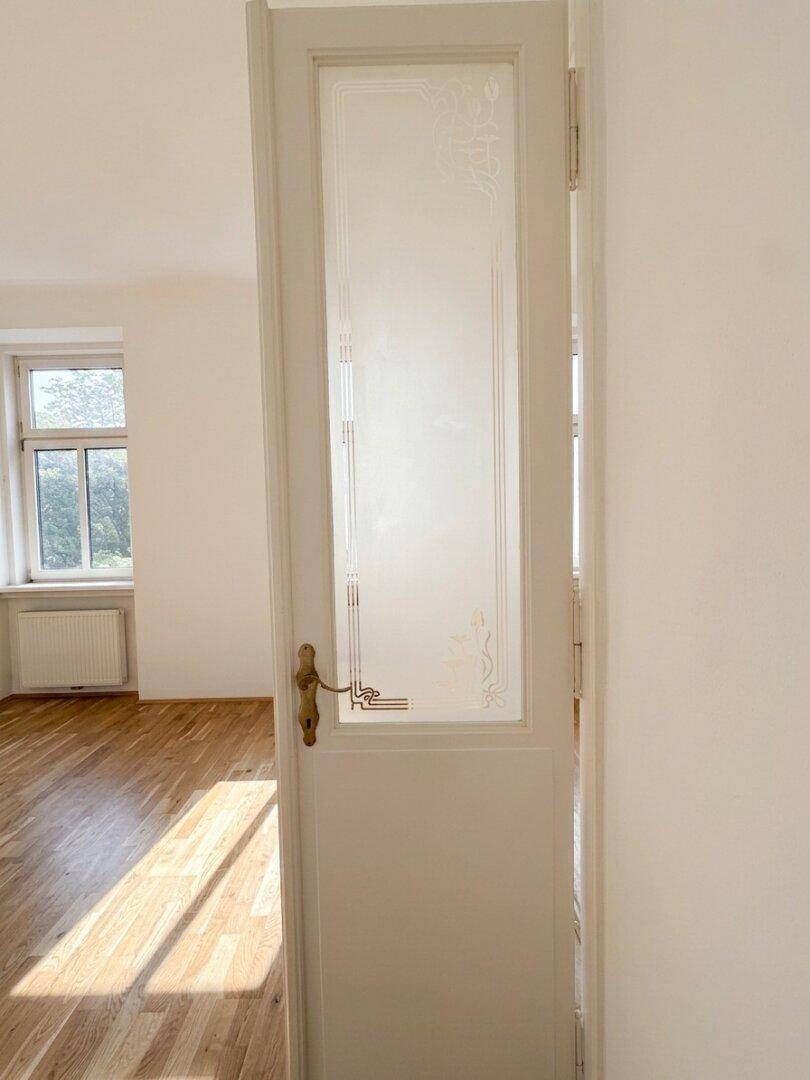 Eine helle Tür mit Glaseinsatz und dekorativem Muster führt in einen sonnigen Raum mit Parkettboden.