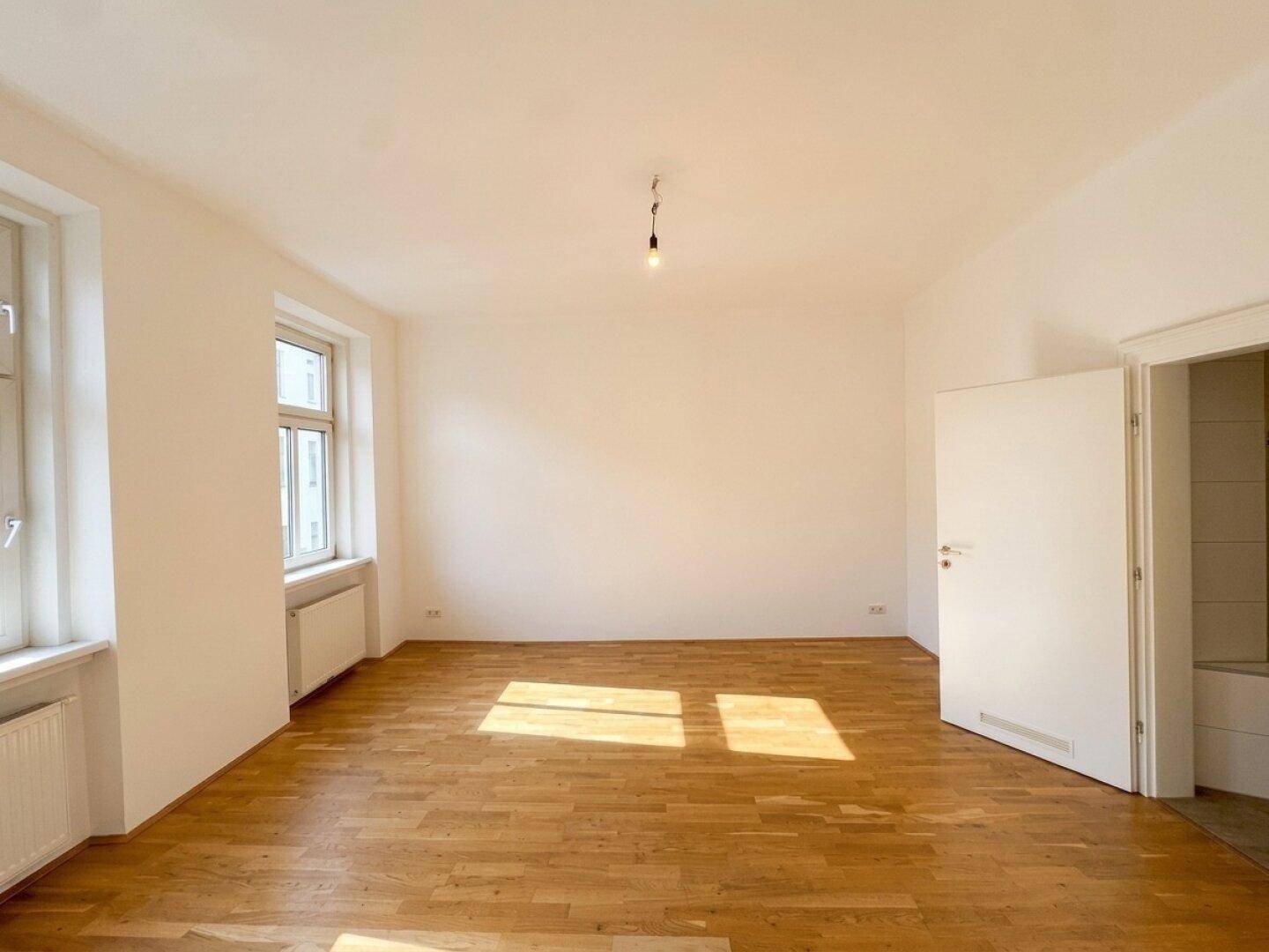 Unmöbliertes Schlafzimmer mit hellem Parkettboden und Fenstern, die den Raum mit Sonnenlicht füllen.