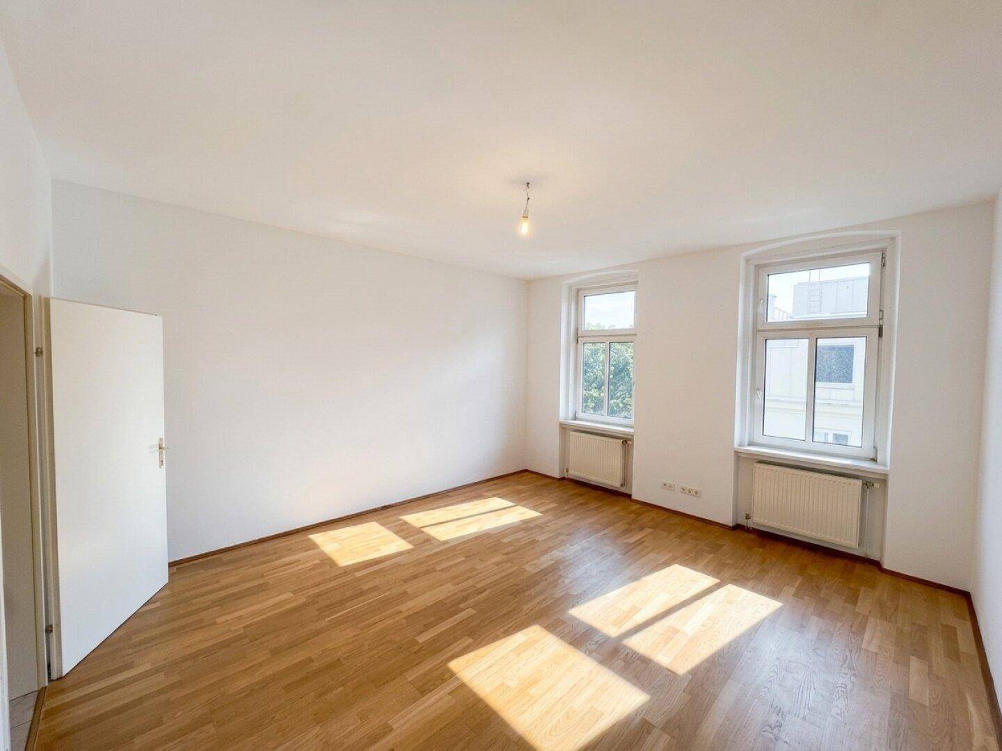 Leeres, helles Zimmer mit Holzboden und zwei großen Fenstern, bereit zur individuellen Gestaltung.