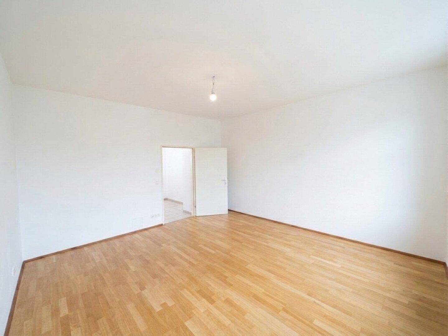 Geräumiges, leeres Zimmer mit Holzboden und offenem Durchgang zu einem weiteren Raum.