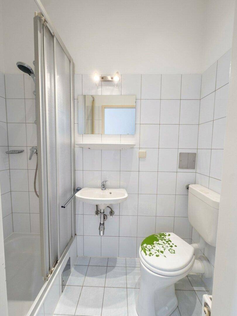 Kleines Badezimmer mit Dusche, Waschbecken und WC, komplett gefliest in Weiß und Hellgrau.