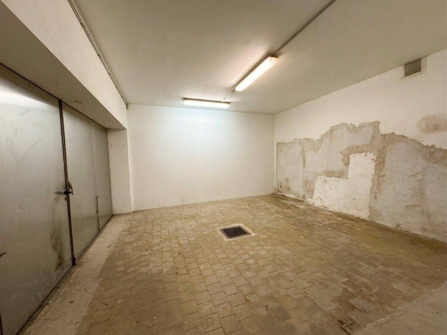 Leerer Garagenraum mit Fliesenboden, weißen Wänden und einer großen Metalltür.