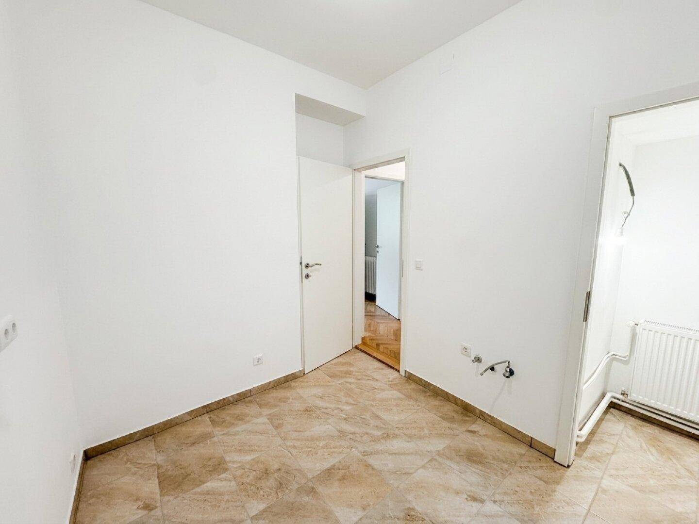 Heller Raum mit Fliesenboden und sichtbaren Anschlüssen für eine zukünftige Küche.