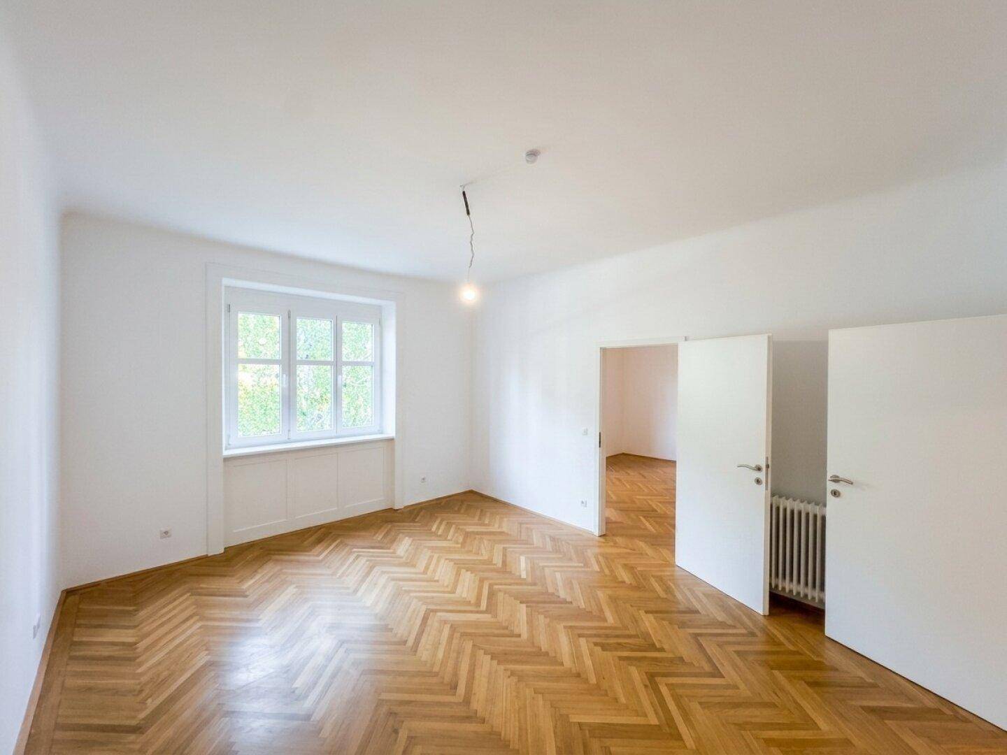 Geräumiges, leeres Wohnzimmer mit Fischgrätparkett, hohen Decken und viel Tageslicht.