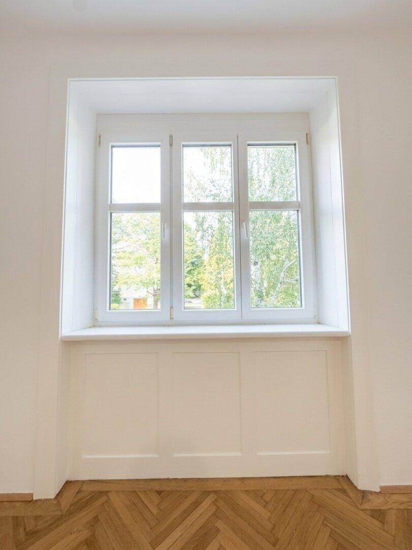 Großes Fenster mit Blick ins Grüne und weißer Holzverkleidung, das viel Tageslicht hereinlässt.