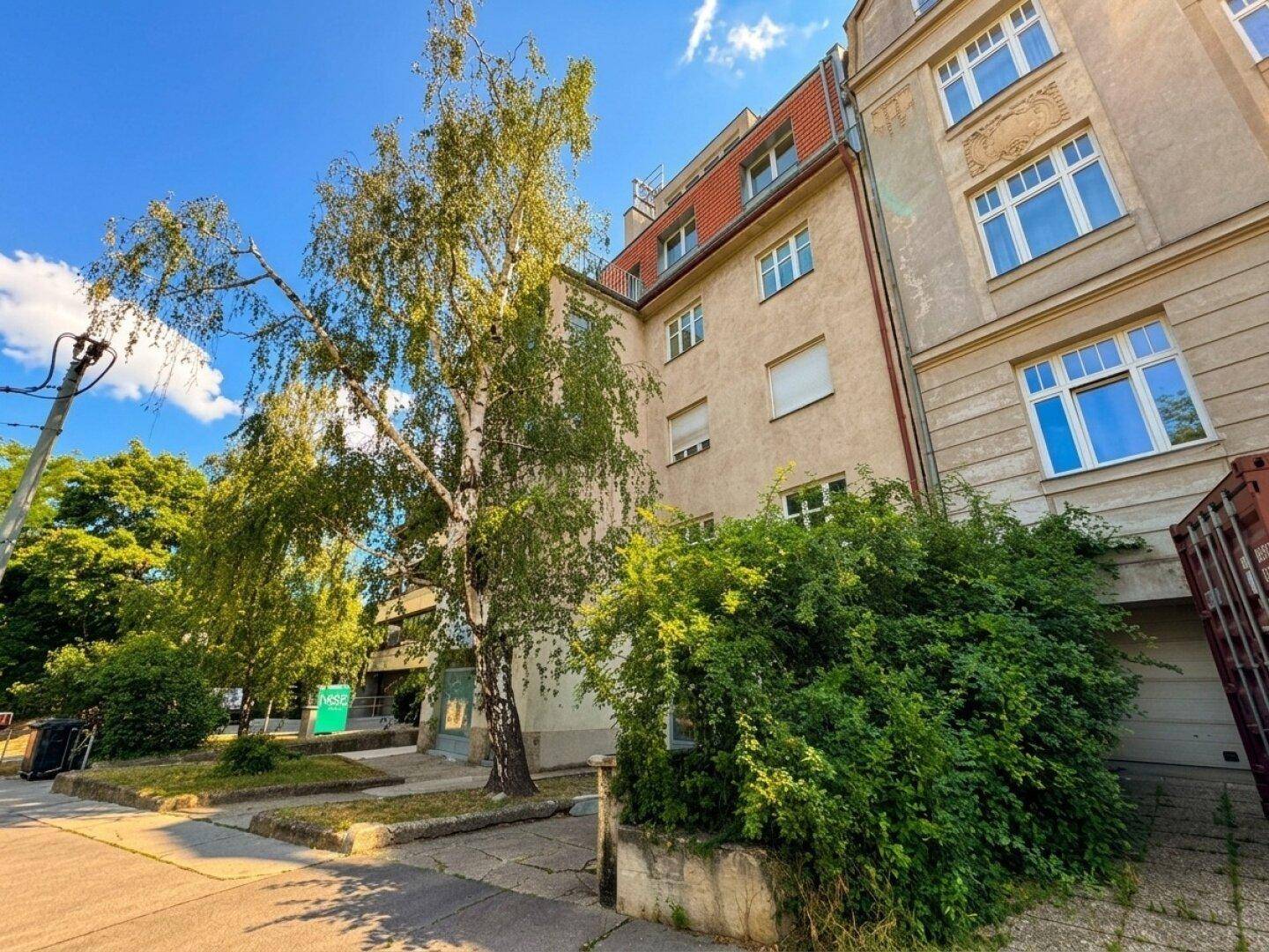 Ansicht des Gebäudes von der Seite mit Bäumen und Büschen entlang der Straße.