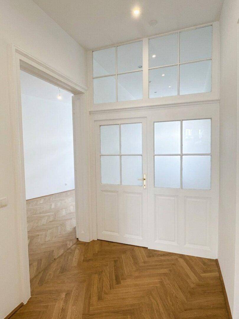 Heller Flur mit Fischgrätparkett und einer eleganten Tür mit Oberlichtfenstern.