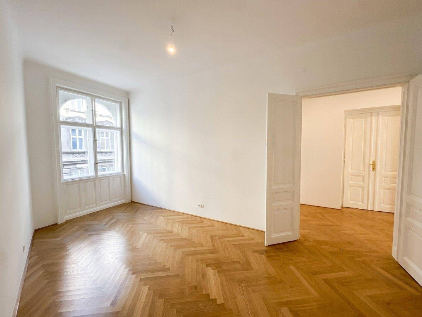 Geräumiges, helles Zimmer mit Fischgrätparkett und hohen Fenstern, bereit zur Einrichtung.