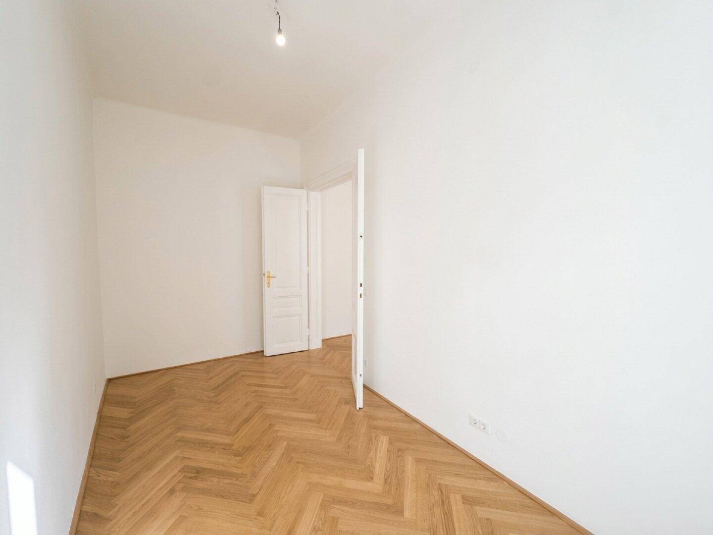 Helles, leeres Zimmer mit Fischgrätparkett und einer offenen Tür zu einem weiteren Raum.