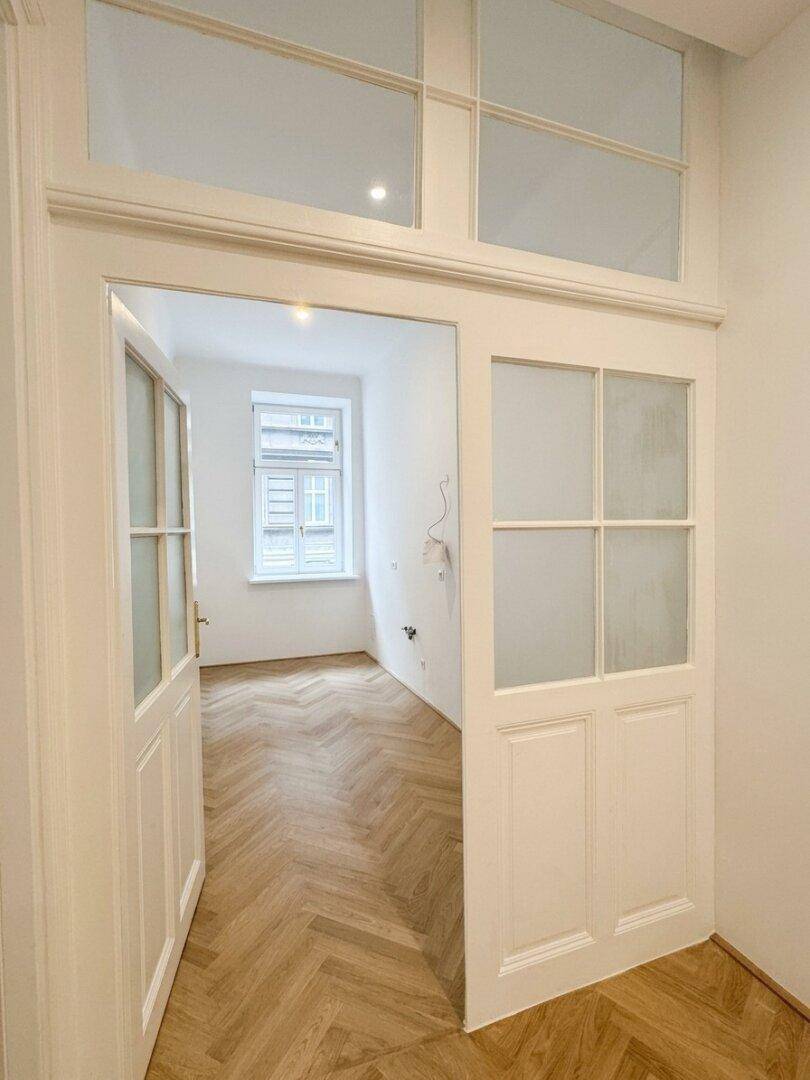 Blick durch eine Tür mit Oberlichtfenstern in einen hellen Raum mit Fischgrätparkett.