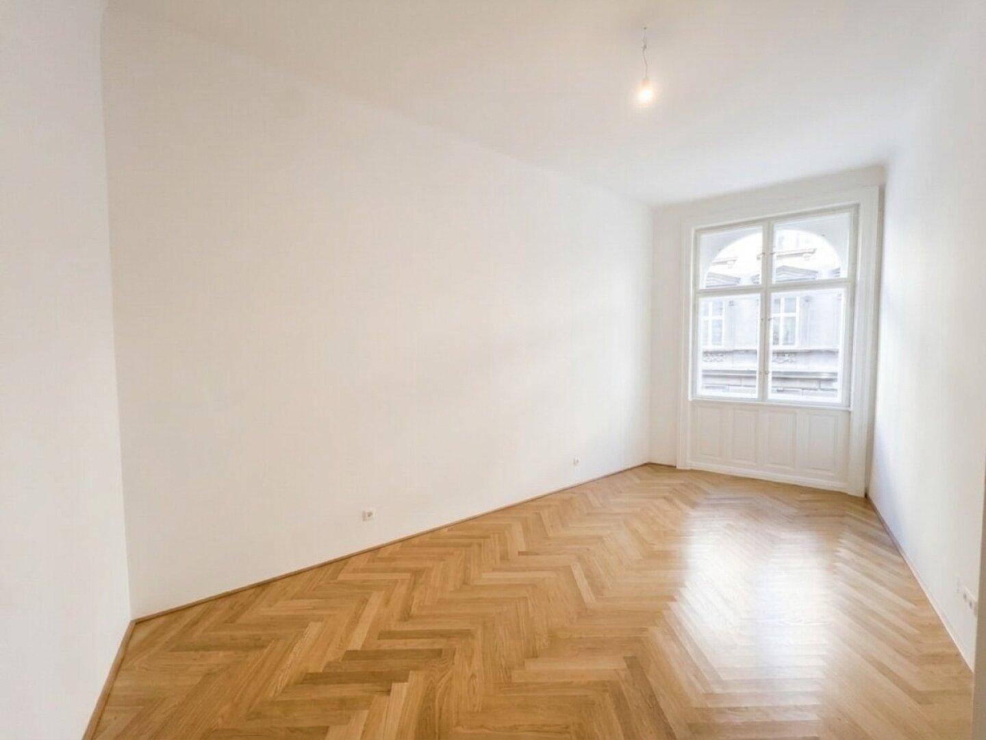 Helles, leeres Zimmer mit Fischgrätparkett und einem hohen Fenster mit Rundbogen.