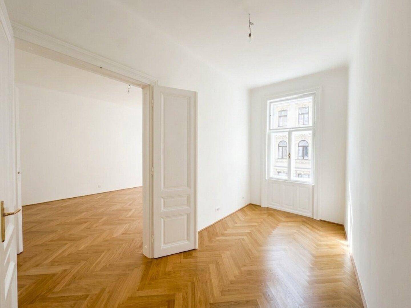 Blick durch eine offene Tür in ein helles Zimmer mit Fischgrätparkett und hohem Fenster.