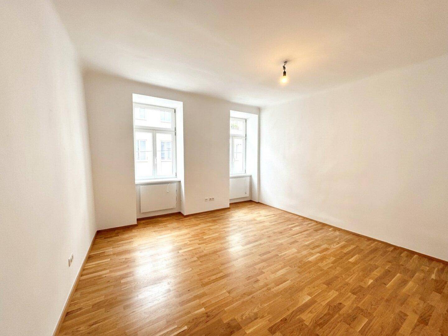 Geräumiges, leeres Zimmer mit Parkettboden und viel Tageslicht durch die Fenster.