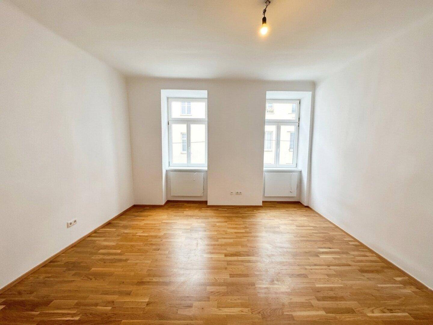 Leeres Zimmer mit hellem Parkettboden und zwei großen Fenstern, bereit zur Einrichtung.